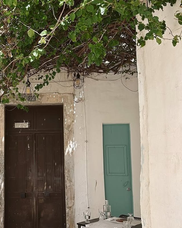 Beco caiado com uma trepadeira por cima, uma porta antiga de madeira, uma porta verde e a ponta de uma mesa posta